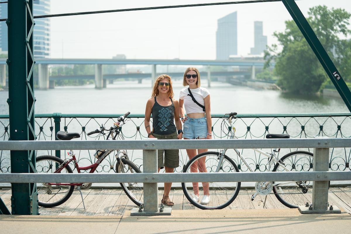 Dr. Lendrum and Arabella Cummings stand on a bridge in Grand Rapids with their bikes, the river and cityscape in the background